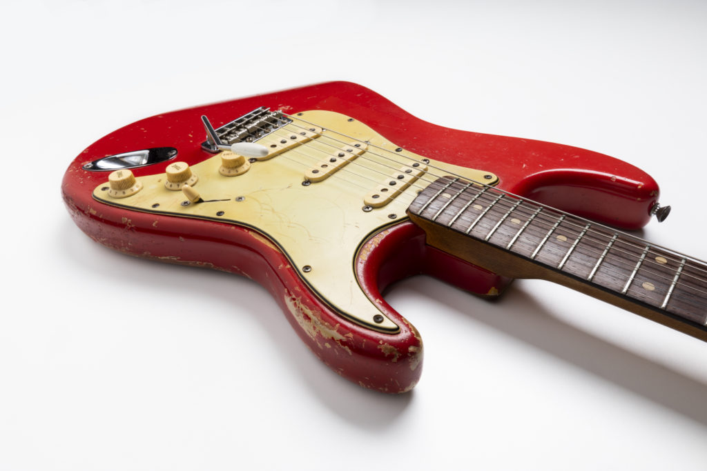 Electric guitar with ‘Dakota red’ body and cream pick-guard. The fret-board is dark rosewood with dot inlays. There are three single-coil pickups with a three-way switch, one volume and two tone control dials. The body has severe road-wear.